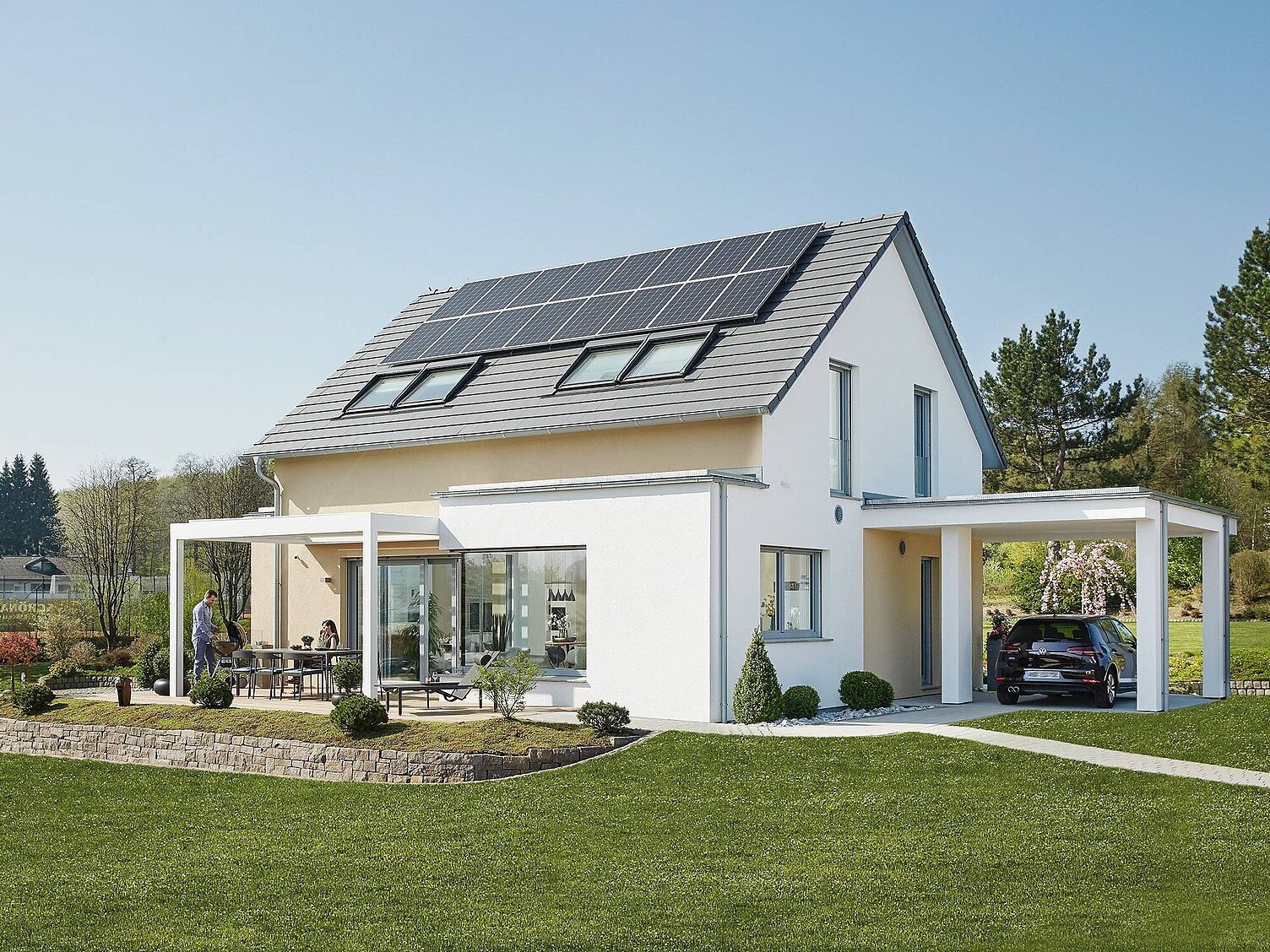 Ein modernes, weißes Einfamilienhaus mit Solaranlagen auf dem Dach. Vor der Terrasse sitzen zwei Personen an einem Tisch. Die Umgebung ist gut gepflegt mit Rasen und kleinen Büschen. Ein schwarzes Auto steht unter einem Carport. Ein klarer blauer Himmel ist im Hintergrund zu sehen.