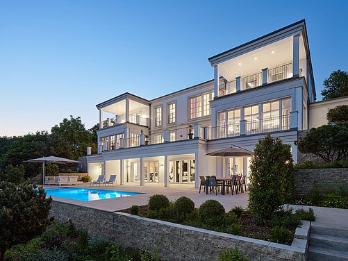 Eine moderne Villa mit mehreren Etagen, großen Fenstern und einem Swimmingpool im Vordergrund. Auf der Terrasse stehen Tische und Stühle, umgeben von Pflanzen und einem Garten. Der Himmel ist blau, es ist Abenddämmerung.