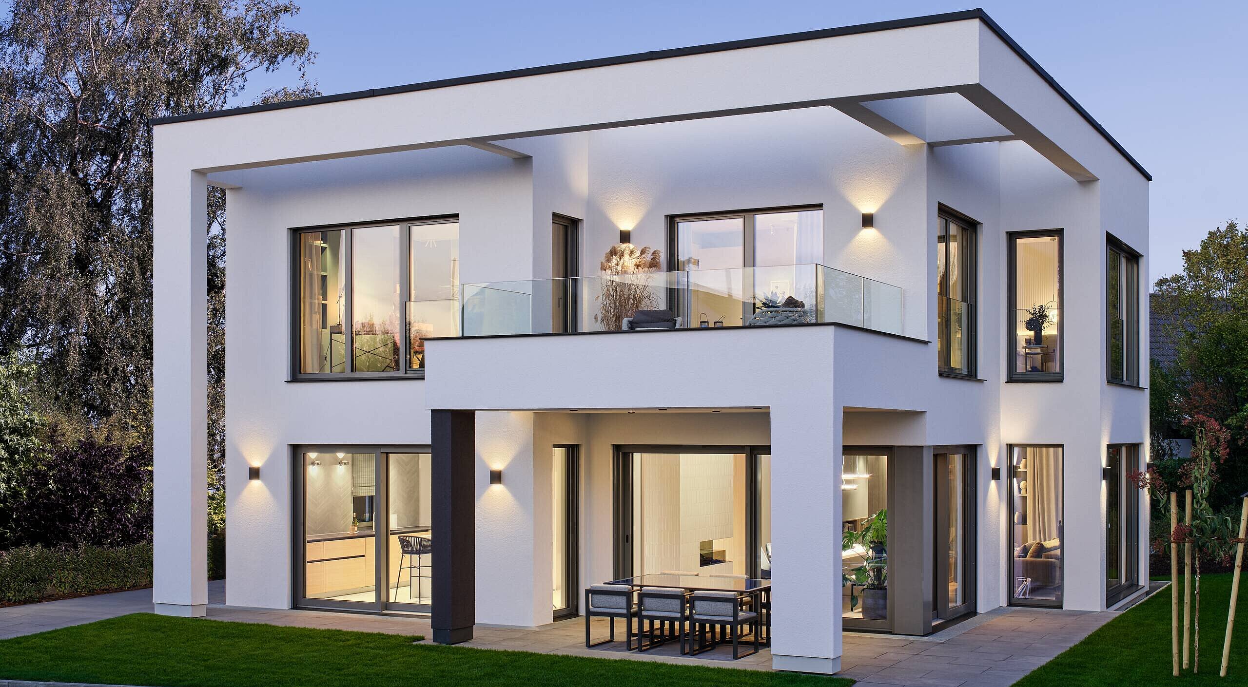 Ein modernes, zweigeschossiges Wohnhaus mit flachem Dach. Es hat große Fenster, die Licht hereinlassen, und einen Balkon. Die Fassade ist hell und schlicht, umgeben von einem gepflegten Garten mit Rasen und wenigen Pflanzen. Der Himmel ist leicht blau, was eine angenehme Abendstimmung vermittelt.