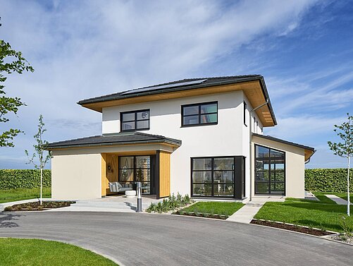 Energieeffizientes Fertighaus von WeberHaus Ein modernes, zweistöckiges Haus mit einem flachen Dach und großen Fenstern. Die Fassade ist überwiegend weiß mit Holzdetails. Im Vordergrund befinden sich ein gepflasterter Weg und ein Rasen, umgeben von kleineren Bäumen und Hecken. Der Himmel ist klar und blau.